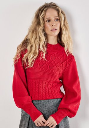 Maglione rosso a maglia con texture a motivo, maniche lunghe a sbuffo e scollatura a coste. Abbinato a una gonna grigia.