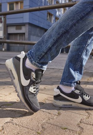 Osoba nosząca czarno-białe sneakersy Nike z widocznymi podeszwami powietrznymi oraz niebieskie dżinsy, stąpająca po wyłożonym chodniku na zewnątrz.