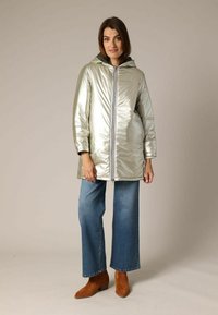 Doudoune métallique argentée avec capuche, fermeture éclair à l'avant et poches latérales, portée sur un jean large bleu et des bottines marron.