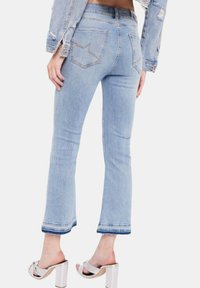 Jeans in denim azzurro chiaro con taglio a campana, dettagli leggermente strappati e un motivo cucito sulle tasche posteriori. L'orlo presenta un accento blu.