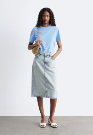 Kvinde med krøllet hår iført lyseblå t-shirt, midnats-lang denim nederdel, hvide åbne sko og bærende en tan skuldertaske, står.