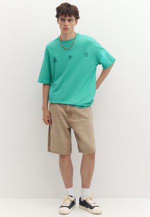 Jeune homme debout portant un t-shirt turquoise oversize, un short beige longueur genoux, des chaussettes blanches et des baskets noires et blanches.