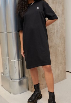 Personne portant une robe T-shirt noire Adidas, des bottes noires à lacets et un collier chaîne en argent, se tenant près de tuyaux métalliques et d'un mur en béton.