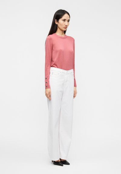 Πουλόβερ με μακριά μανίκια σε ροζ απόχρωση ροζ, με διακοσμητικά κουμπιά στις μανσέτες· συνδυασμένο με λευκό, ψηλόμεσο, παντελόνι με φαρδύ πόδι και μαύρα παπούτσια με μυτερή μύτη.