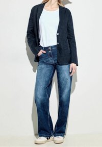 Námořnický lněný blazer, bílý tričko a modré džíny s rozšířenými nohavicemi, doplněné bílými teniskami, stojící na světlém pozadí.