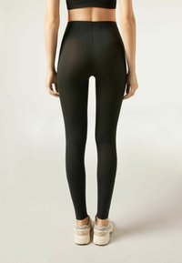 Femme portant des leggings noirs taille haute et des baskets blanches, se tenant avec les mains détendues sur les côtés devant un fond clair uniforme.