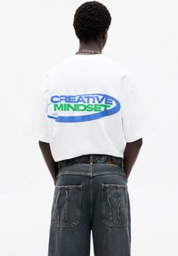White cotton t-shirt, oversized fit, features "CREATIVE MINDSET" in blue and green print on the back, paired with dark denim shorts.