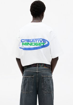 White cotton t-shirt, oversized fit, features "CREATIVE MINDSET" in blue and green print on the back, paired with dark denim shorts.