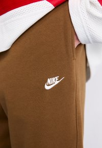 Person iført brune Nike sweatpants med hvidt logo, hånd delvist i lommen, og en hvid og rød mesh-top synlig over bukserne.