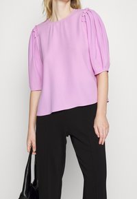 Blusa lilla con maniche a sbuffo corte e dettagli plissettati sulle spalle. Indossata con pantaloni neri, caratterizzati da una texture liscia e morbida.