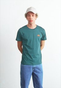 Zelena bombažna majica s krožnim izrezom in oranžnim logotipom na levem prsnem delu, v kombinaciji z modrimi denim hlačami in belo kapo.