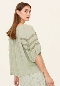 Hellgrüne Bluse mit Puffärmeln, die mit Spitzenstreifen verziert ist. Strukturiertes Material mit lässiger Passform, kombiniert mit einem Rock im Blumenmuster.