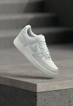 Pantof sport alb Nike Air Force 1 cu panouri din plasă și piele, șireturi legate, plutind deasupra unei margini de beton cu un fundal neclar de trepte.