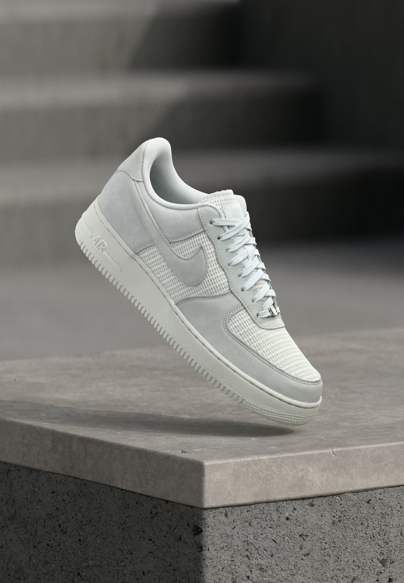 Pantof sport alb Nike Air Force 1 cu panouri din plasă și piele, șireturi legate, plutind deasupra unei margini de beton cu un fundal neclar de trepte.