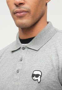 Γκρι πόλο μπλουζάκι με γιακα, διαθέτοντας ένα μαύρο και λευκό ραμμένο πατς με μια φιγούρα σε γυαλιά ηλίου στην αριστερή πλευρά.