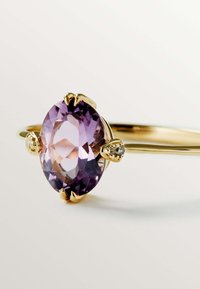 Anillo de oro con una gema ovalada de color púrpura, rodeada de pequeños diamantes a cada lado; acabado suave y textura pulida.