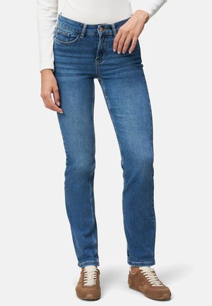 Jean en denim bleu avec une coupe droite, présentant un design à cinq poches et une fermeture à bouton, portés avec des baskets marron et un haut à manches longues blanc.