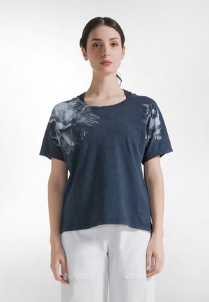 Kvinde med mørkt hår bundet tilbage iført en marineblå t-shirt med grafik og hvide bukser, stående foran en ensartet lys baggrund.