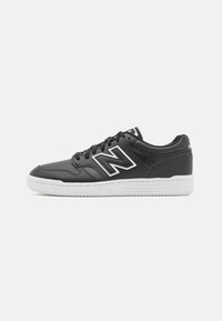 Svarta lädersneakers med vit gummisula, med en stor silverfärgad "N"-logotyp, rund tå och svarta snören. Slät textur, avslappnad design.