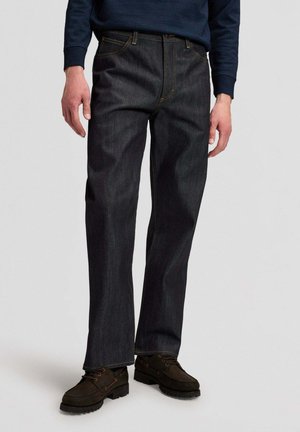 Mann trägt dunkelblaue Jeans mit geradem Bein, dunkelbraune Schnürschuhe und ein marineblaues Langarmhemd vor einem einfarbigen Hintergrund.