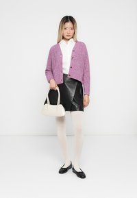 Kvinne iført en rosa knappet cardigan, hvit skjorte, svart skinnskjørt, hvite strømpebukser, svarte leiligheter og holder en hvit håndveske.