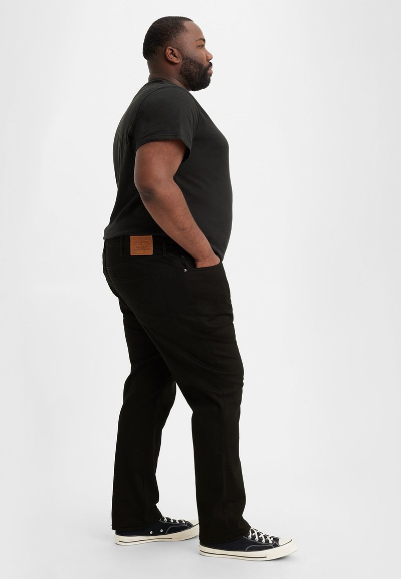 Pantalon en denim noir avec une coupe droite, poches latérales et une étiquette en cuir beige au niveau de la taille. Porté avec un t-shirt noir et des baskets.