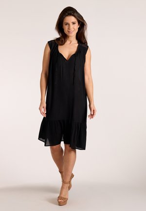 Žena kráčajúca dopredu v čiernych šatách bez rukávov s volánovým lemom a hnedých vysokých pletených sandáloch na jednoliatom svetlom pozadí.