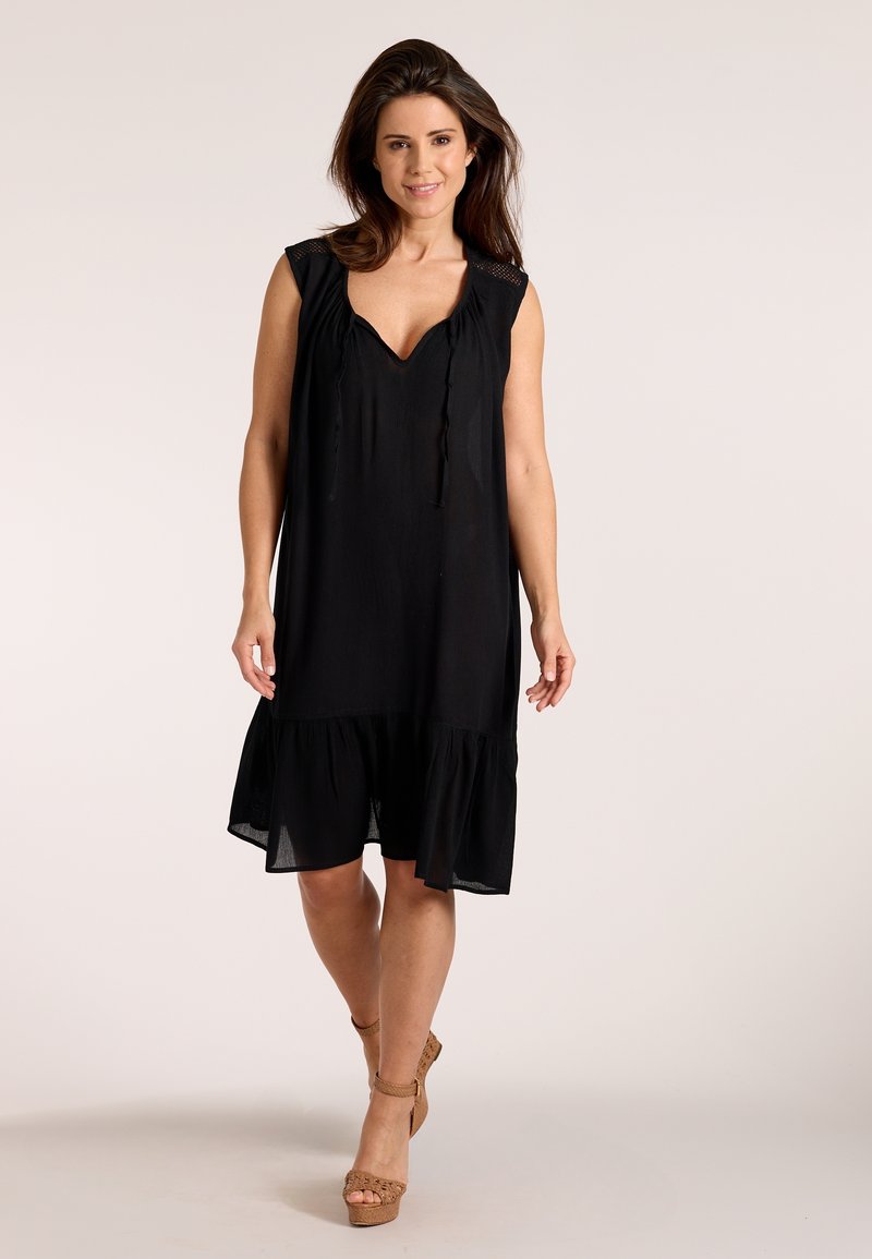 Ženska hodi naprej, oblečena v črno obleko brez rokavov z naborki na spodnjem robu in rjavimi tkanimi sandalmi z visokimi petami, na enostavni svetli podlagi.