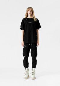 Camiseta negra de gran tamaño con un parche central. Pantalones joggers negros con acentos gráficos en blanco y cremalleras. Terminados con botas blancas de cordones.
