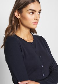 Tumedat sinine kardigan ümmarguse kaelusega, pikkade varrukatega ja viie nupuga kinnitusega. Pehme tekstuur ja keha lähedane disain. Õrn ribakujuline detail.