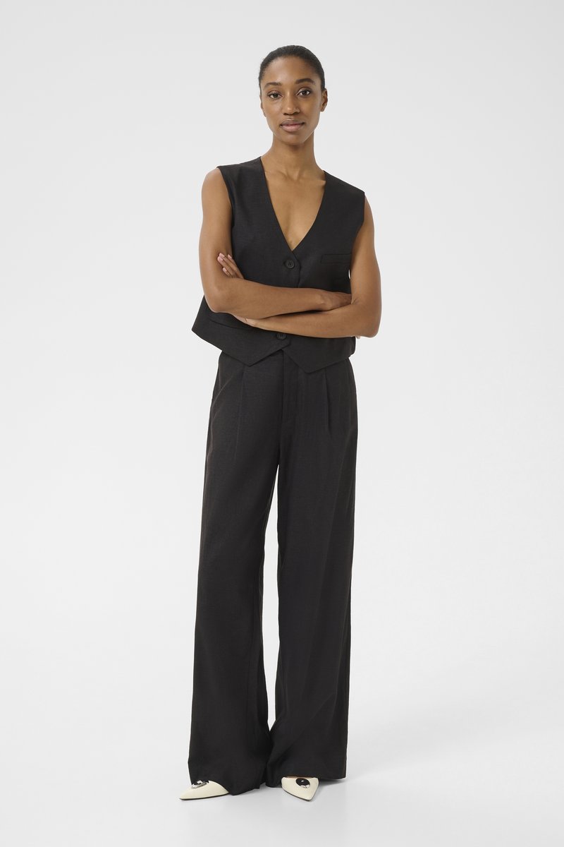 Czarna kamizelka z głębokim dekoltem w kształcie V i pojedynczym guzikiem, w połączeniu z czarnymi spodniami w wysokim stanie. Wąskie białe buty z czarnymi akcentami.