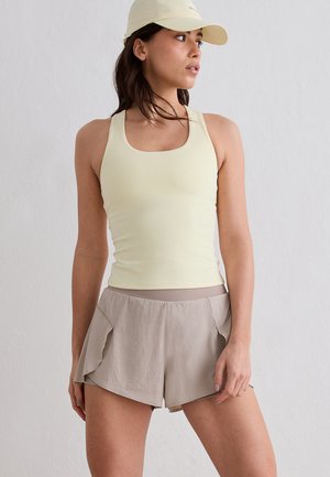 Woman wearing a light yellow tank top, beige athletic shorts with ruffled sides, and a matching light yellow baseball cap, standing against a white wall.