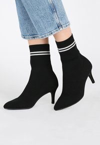 Czarne botki za kostkę o dzianinowej fakturze, o spiczastych czubkach i 5-centymetrowych obcasach w stylu stiletto. Posiadają prążkowany mankiet z dwoma białymi paskami.