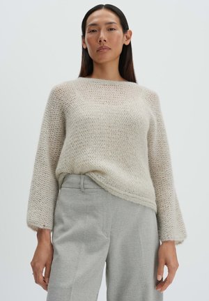 Woman wearing a loose, cream mohair sweater and light gray high-waisted trousers, standing against a plain light background.