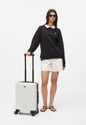 Valise rigide blanche avec texture rainurée, poignée rétractable noire et roulettes ; accompagnée d'un sweat-shirt noir et d'un short blanc.