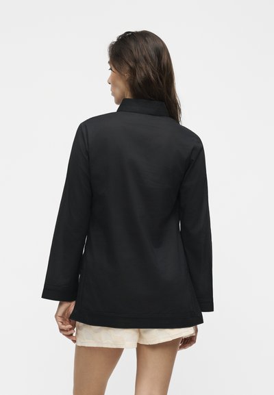 Γυναίκα με μακριά σκουρόχρωμα μαλλιά, που φοράει μαύρο μακρυμάνικο μπουφάν και ανοιχτόχρωμα σορτς, γυρισμένη πλάτη προς ένα απλό φόντο.