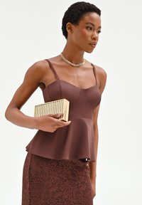 Mujer con una blusa sin mangas marrón con peplum y falda de encaje, sosteniendo un bolso de mano dorado con rayas y llevando un collar grueso de cadena dorada.