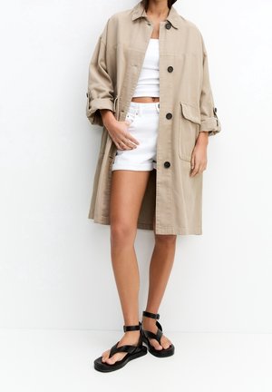 Nainen, jolla on yllään beigenvärinen pitkä takkki valkoisen lyhytyläosan ja shortsejen päällä, seisoo mustat nilkkaremmi-sandaalit jalassaan valkoista taustaa vasten.