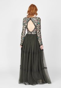 Larga gown de color verde oscuro con adornos florales, mangas largas transparentes y un profundo escote en la espalda. Falda de tul en capas con textura de puntos.