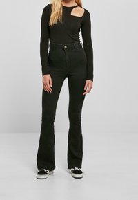 Mujer vestida con pantalones de campana negros de tiro alto, una blusa negra de manga larga con detalle de corte en el hombro y zapatillas en blanco y negro, de pie frente a un fondo liso.