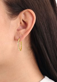 Orecchino a cerchio in oro con design texture, impreziosito da accenti trasparenti. Forma circolare, metallo leggero, indossato nel lobo.