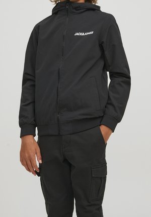 Personne portant une veste à capuche noire zippée avec le logo "JACK & JONES" et un pantalon cargo noir, debout avec une main dans la poche.
