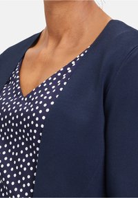 Cardigan bleu marine à finition texturée, porté par-dessus un haut bleu marine à pois blancs, col en V et silhouette ajustée.