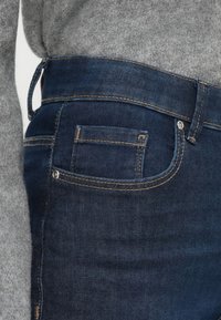 Tamno plave traperice s glatkom teksturom, koje imaju žute šavove, mali stražnji džep i detalj za remen.