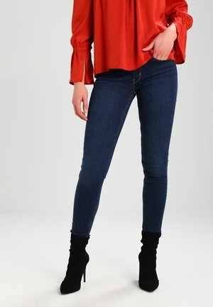 Slim-fit donkerblauwe jeans van denim, met een standaard vijf-pocket ontwerp, gecombineerd met een losse rode blouse en zwarte hoge hakken.