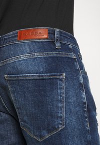 Medium wash denim jeans with a leather Gabba logo patch on the back, two back pockets, and a distressed texture on the fabric.