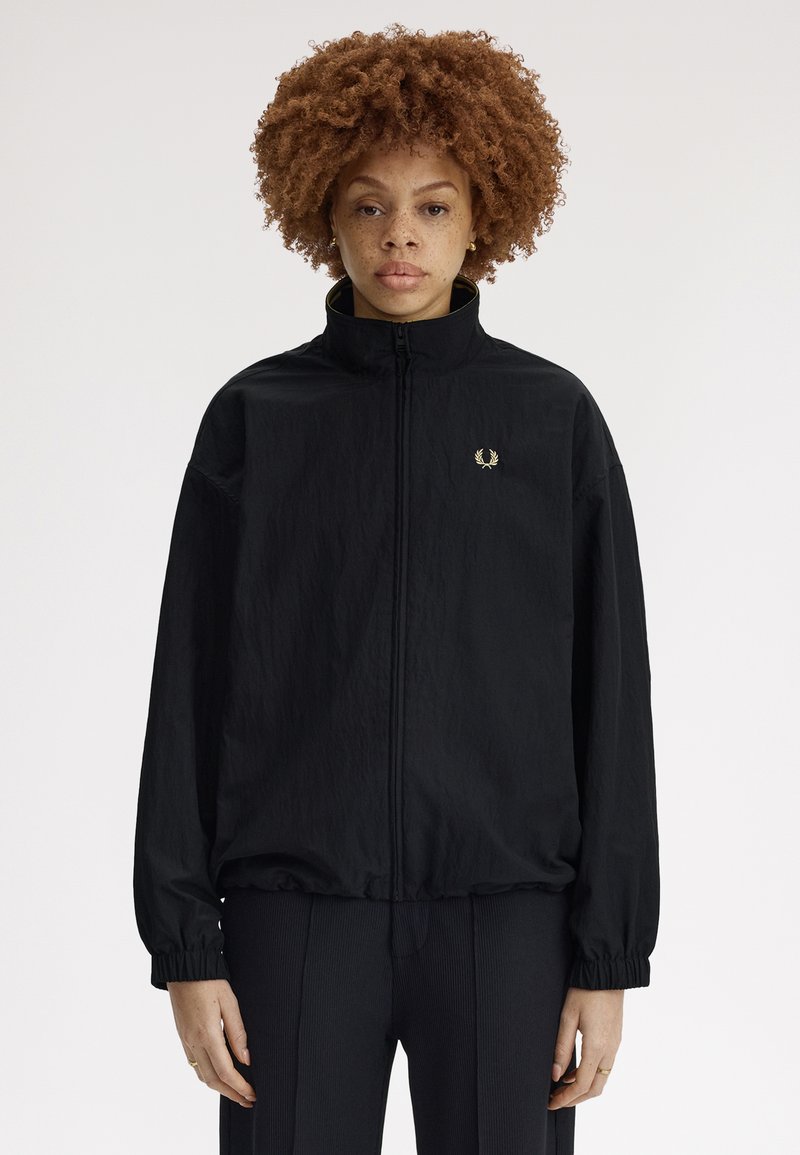 Personne aux cheveux roux bouclés portant une veste noire à fermeture éclair avec des poignets élastiques et un pantalon noir assorti, debout devant un fond uni.