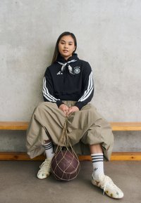 Sudadera negra con rayas y logotipo blanco, pantalones beige oversized y zapatillas con estampado. Pelota marrón en una bolsa de red al lado de una figura sentada.