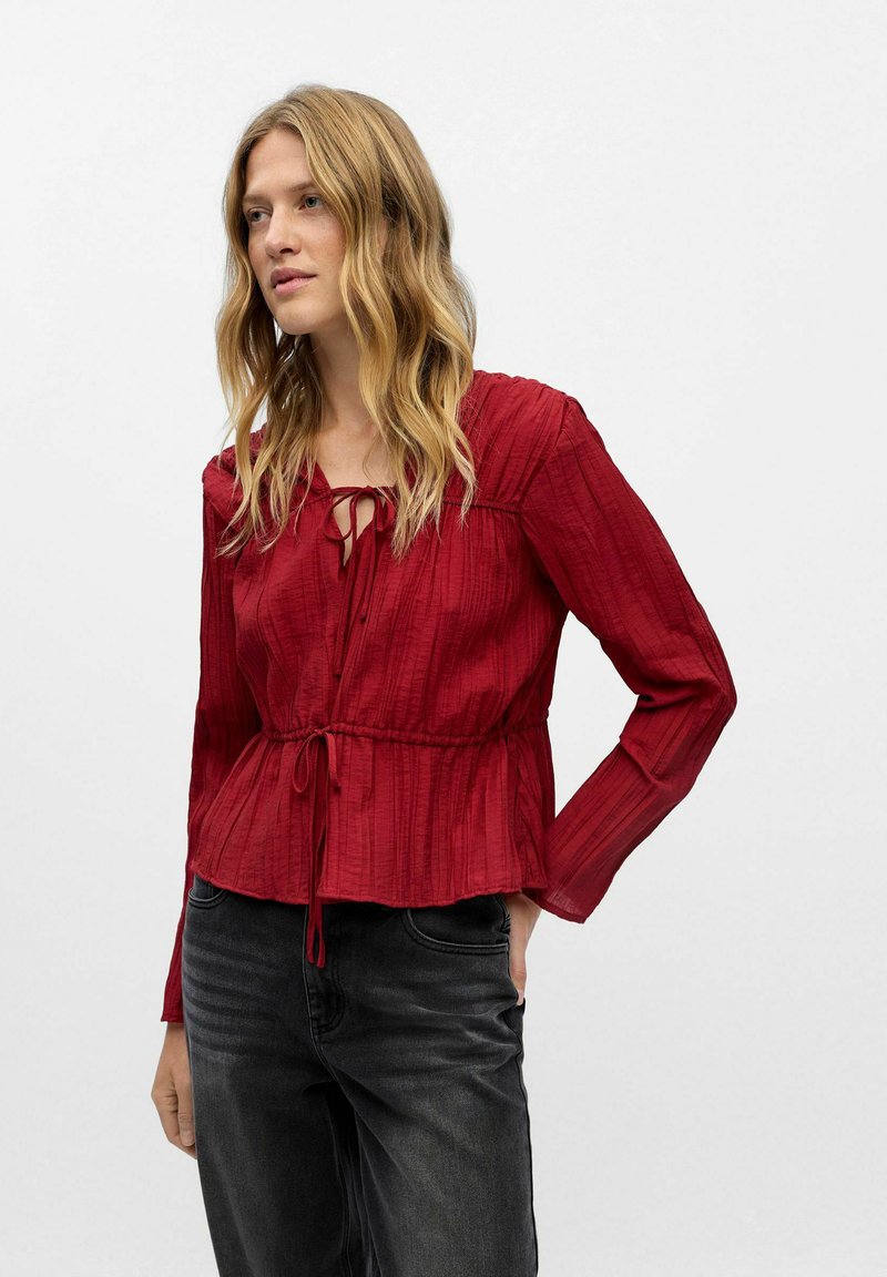 Donna con i capelli biondi lunghi, che indossa una blusa rossa a maniche lunghe con texture e jeans neri, in piedi con una mano in tasca su uno sfondo bianco.
