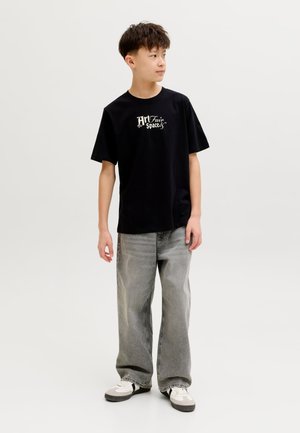 Paauglys berniukas dėvi juodą marškinėlį su grafiniu dizainu, laisvas pilkas džinsus ir baltus sportbačius, žiūri į šoną priešais neutralią foną.